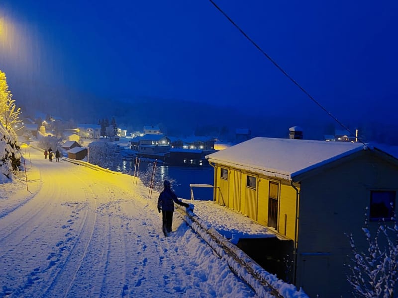 Winterwanderung durch eine verschneite Dorfstraße in Norwegen bei tiefblauer Nachtatmosphäre mit Fjordblick.
