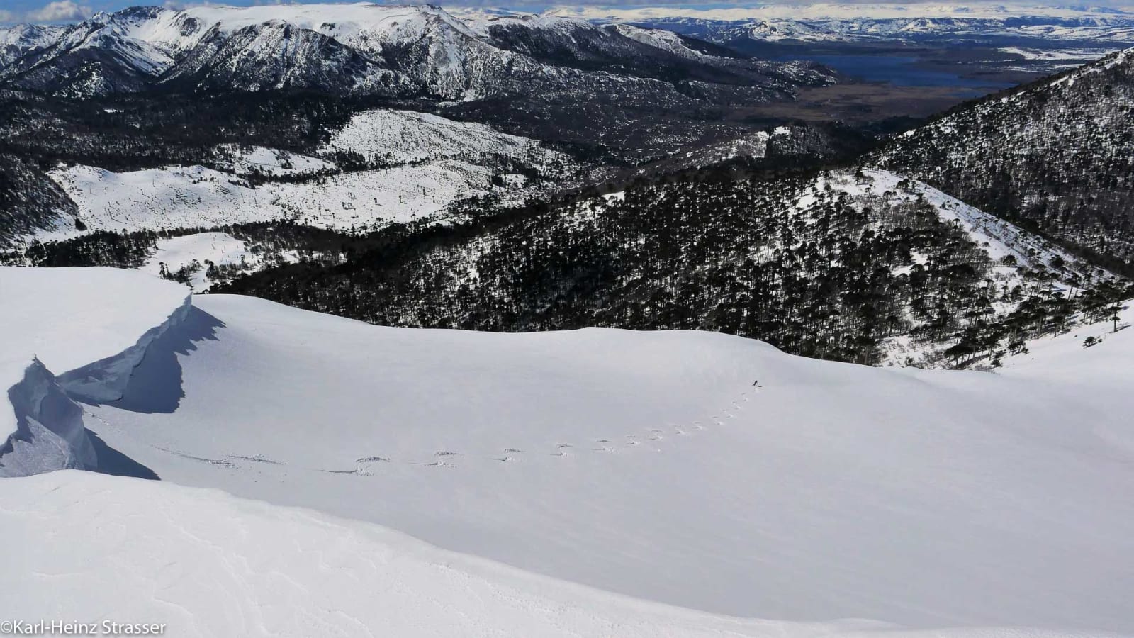 Bergtour Abfahrtsgenuss pur: Einzelne Person zieht Tiefschnee-Spur an sonnigem Hang über verschneitem Tal mit See und Gebirgskette