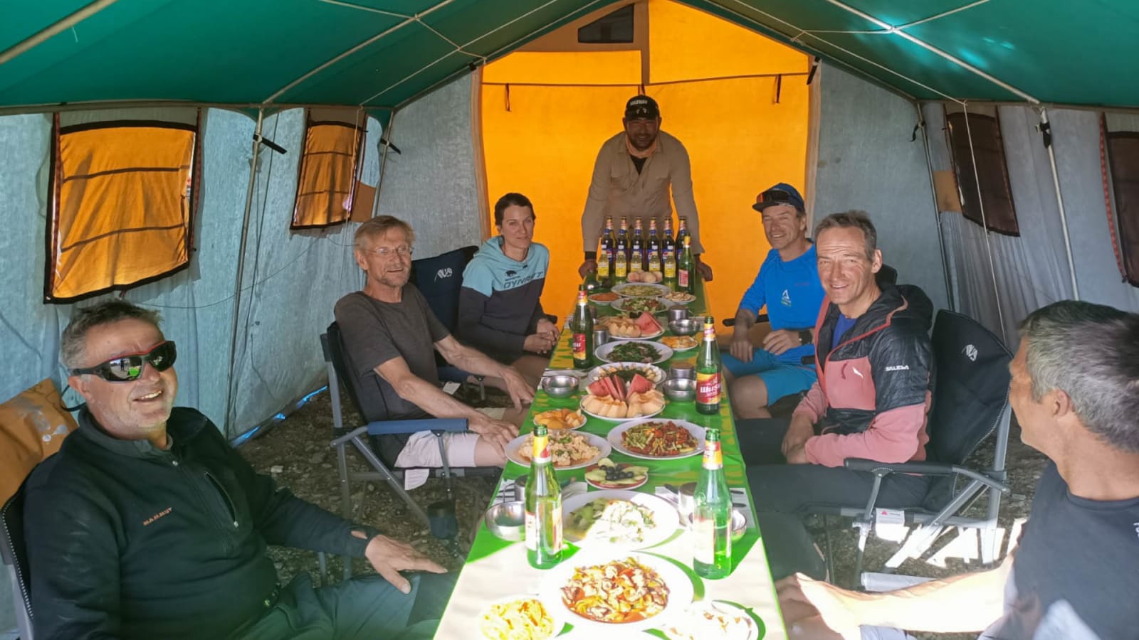 Bergtour-Gruppe bei einem Festmahl im Expeditionszelt nach Gipfelerfolgen, sieben Bergsteiger am gedeckten Tisch.