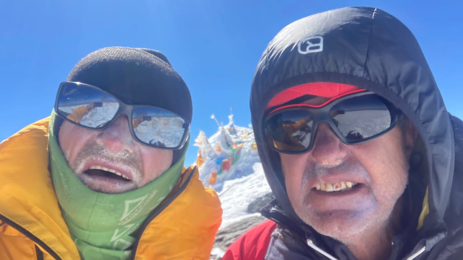 Bergtour Muztagh Ata: zwei Bergsteiger mit Reif im Gesicht vor schneebedecktem Gipfel mit Gebetsfahnen.