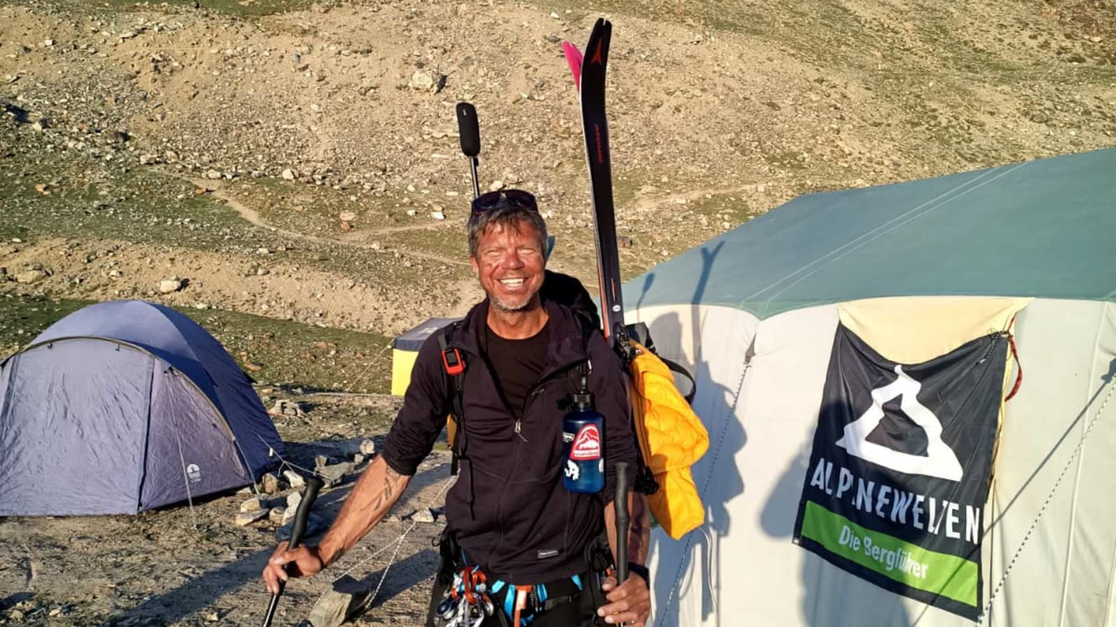 Freudestrahlender Gipfelbesteiger mit Rucksack und Skistöcken vor Zeltlager auf steinigem Gelände bei einer Bergtour.