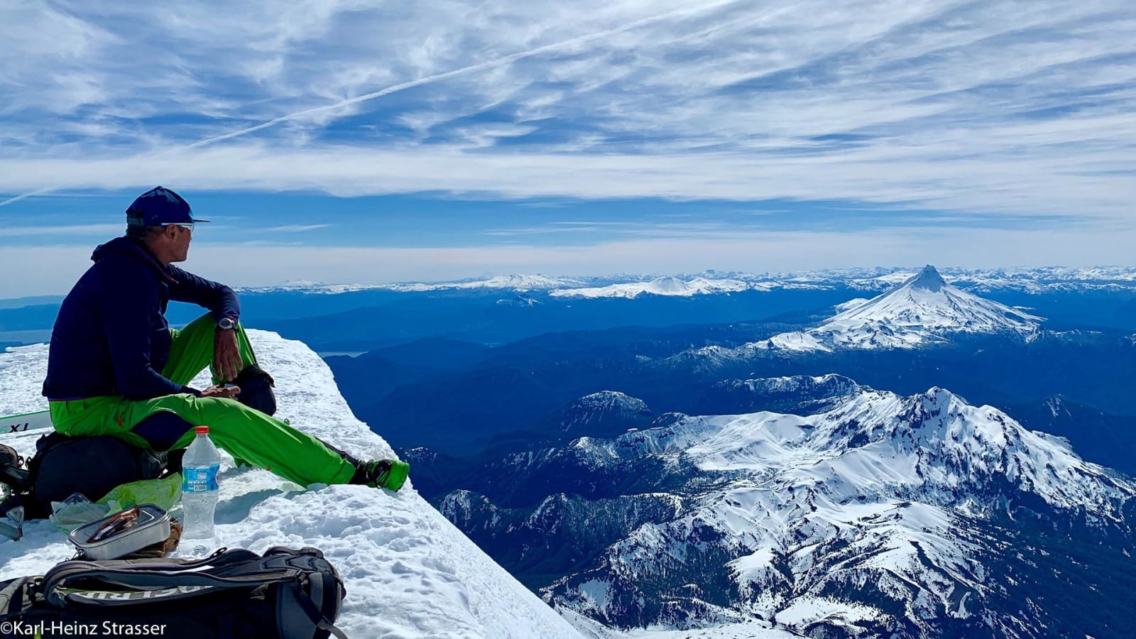 Bergsteiger rastet auf schneebedecktem Gipfelgrat der Aussichtskanzel mit Vulkankegel im Hintergrund.