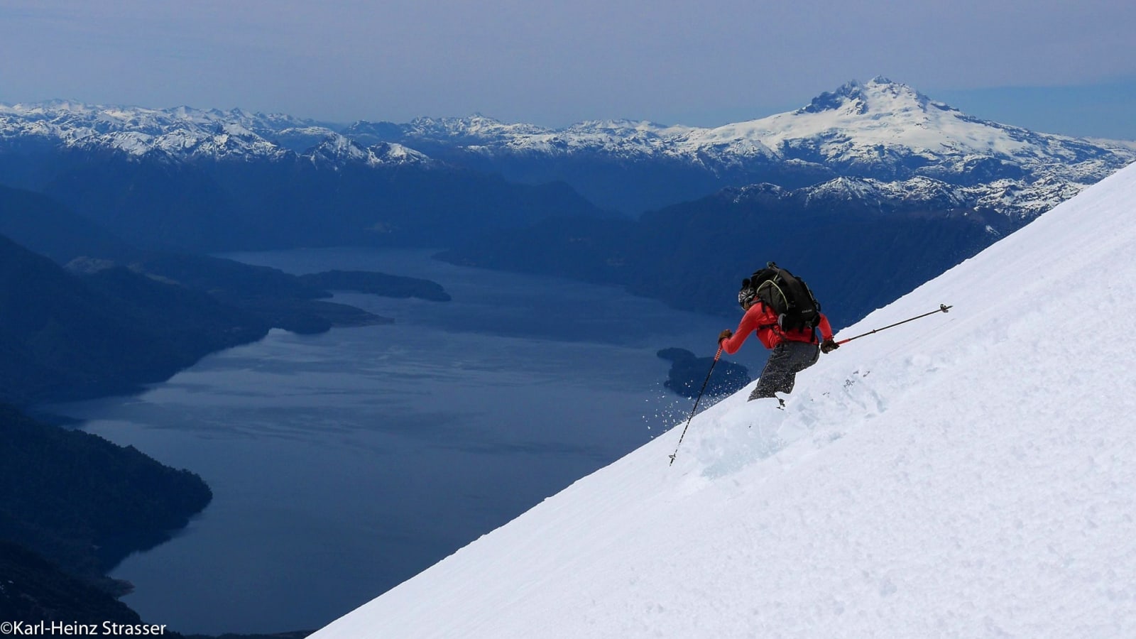 Skitour am Lago Todos los Santos: Bergsteiger in roter Jacke fährt steilen Schneehang mit Seepanorama ab.