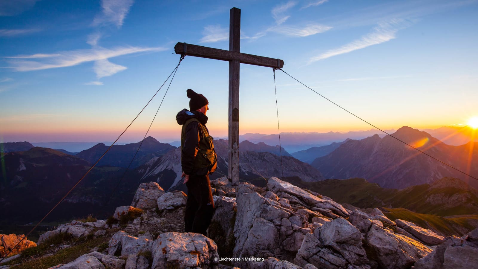 Bergwanderung auf den Augstenberg in Liechtenstein: Wanderer steht beim Gipfelkreuz mit Panorama im Gegenlicht.