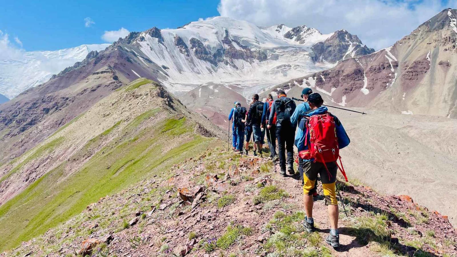 Expedition Pik Lenin: Gruppe von Höhenbergsteigern wandert in Einzeillinie entlang felsigem Grat mit vergletschertem Hochgebirge