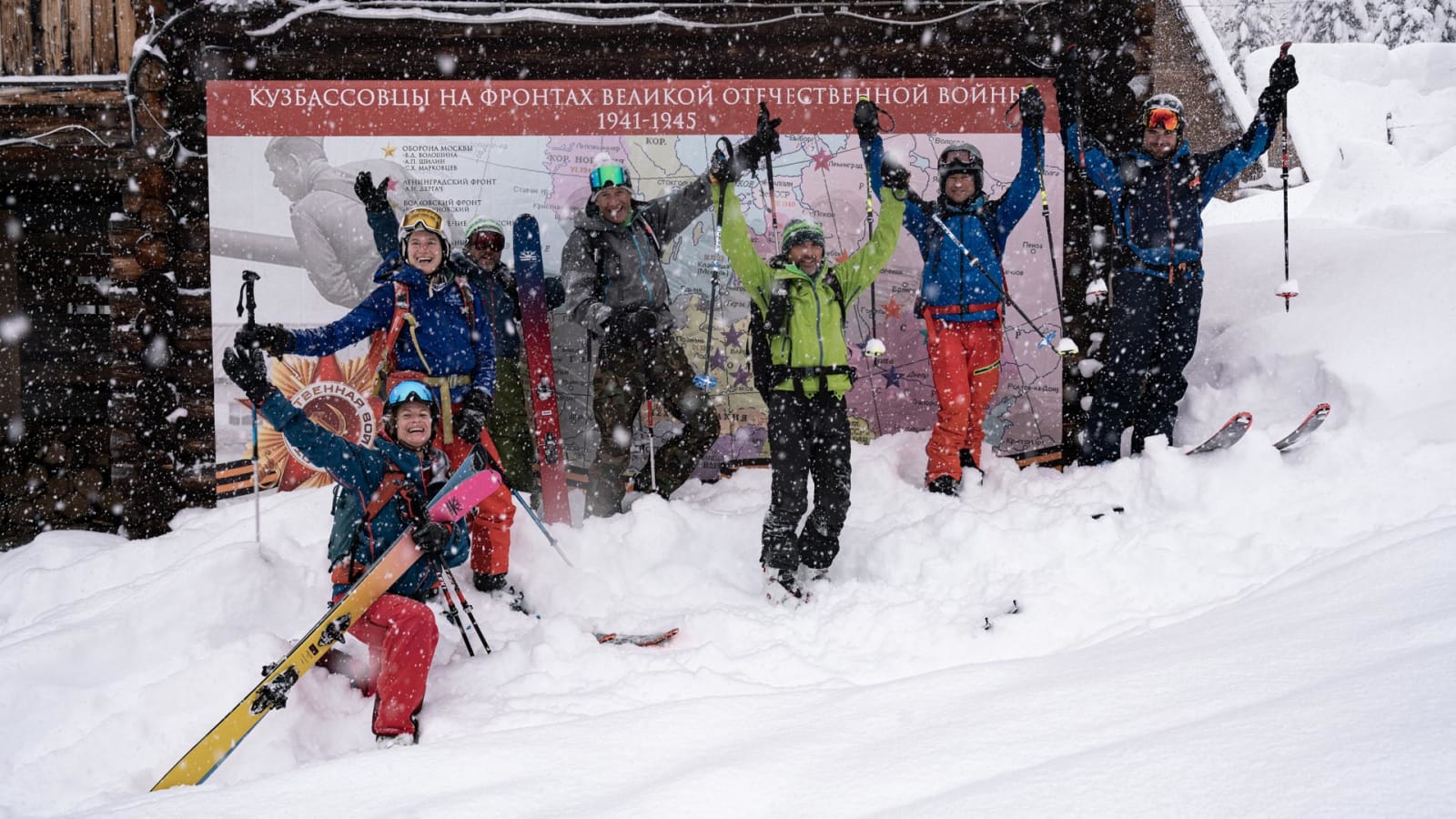 Skitour-Gruppe von sieben Skitourengehern jubelt vor kyrillischer Informationstafel in Luzhba bei Schneefall.