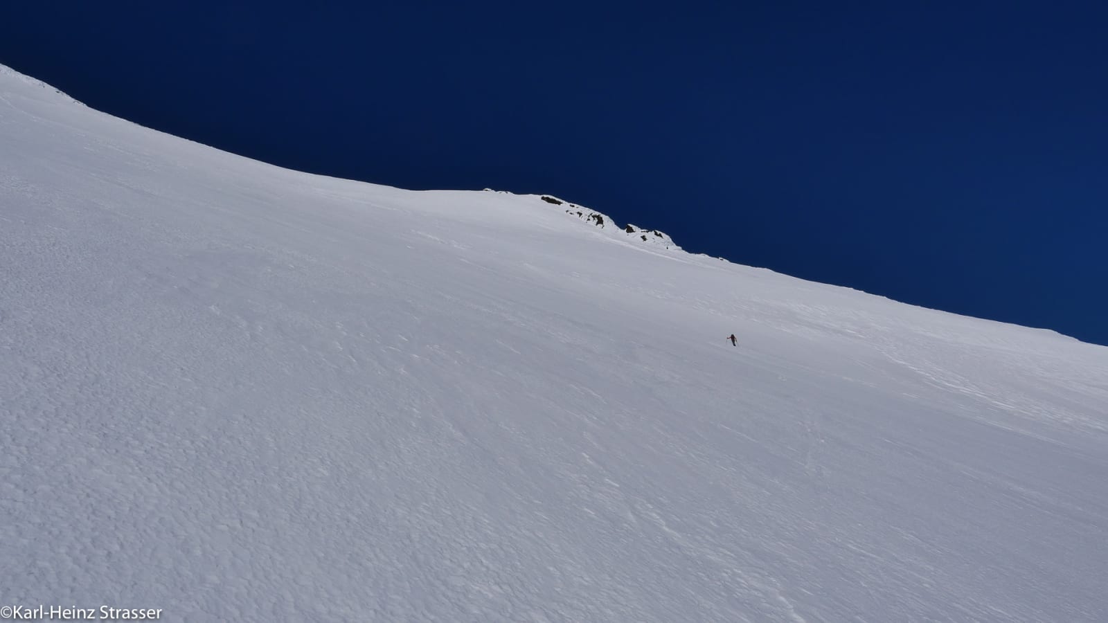 Skitourengeher als Silhouette auf weite unberührte Skitourenhänge bei der Abfahrt unter tiefblauem Winterhimmel – Bergtour