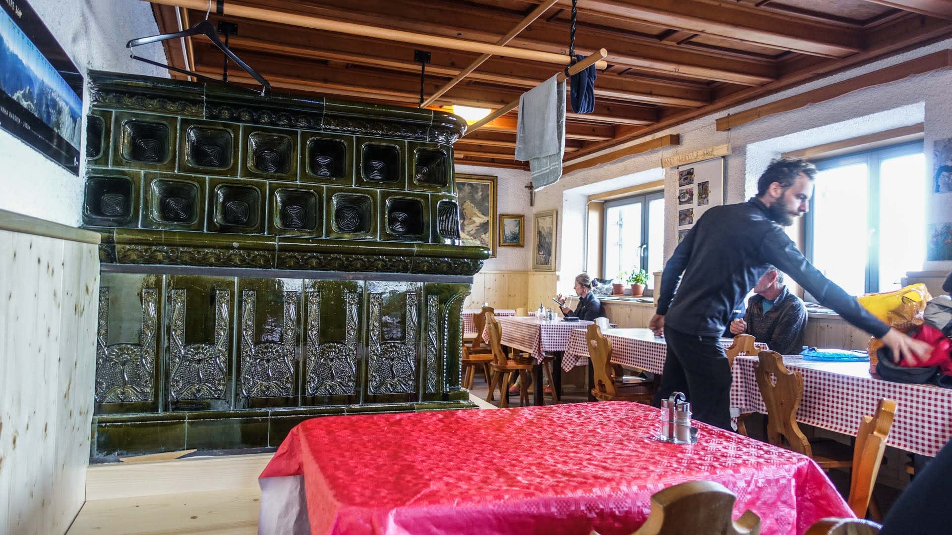 Bergwanderer im Gastraum des Dom na Kriških Podih mit grünem Kachelofen und Holzdecke in Slowenien.