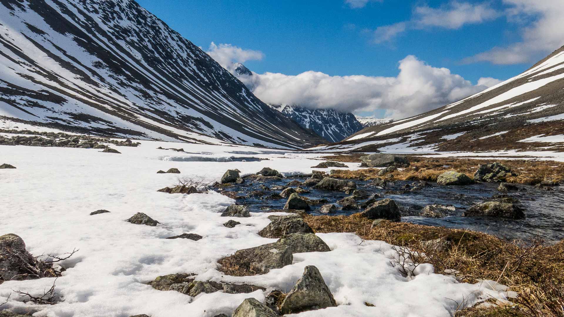 Schneebedecktes U-Tal mit Gebirgsbach und pyramidalem Gipfel bei einer Bergwanderung in den Lyngenalpen.