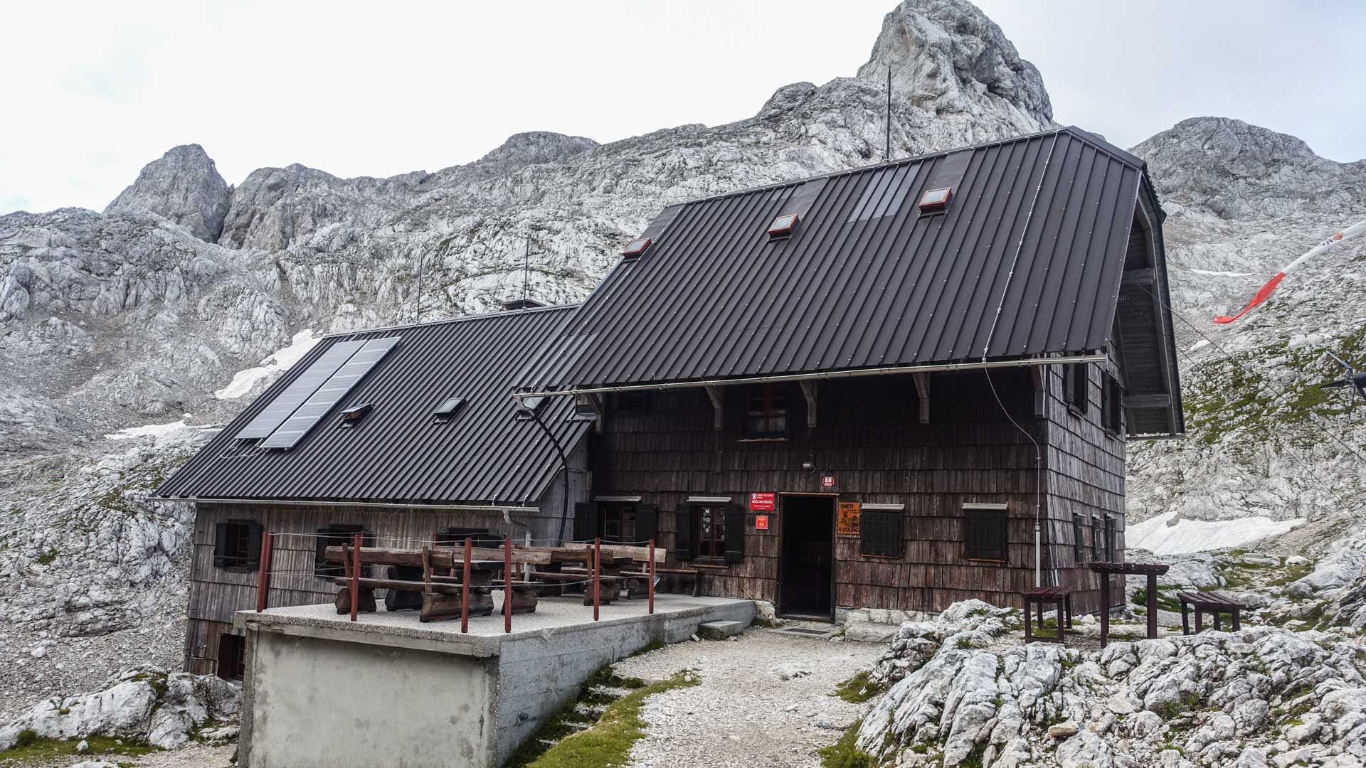 Bergwanderung in Slowenien: Holzberghütte Koča na Dolicu vor verkarsteten Kalkfelsen mit Schneeresten.