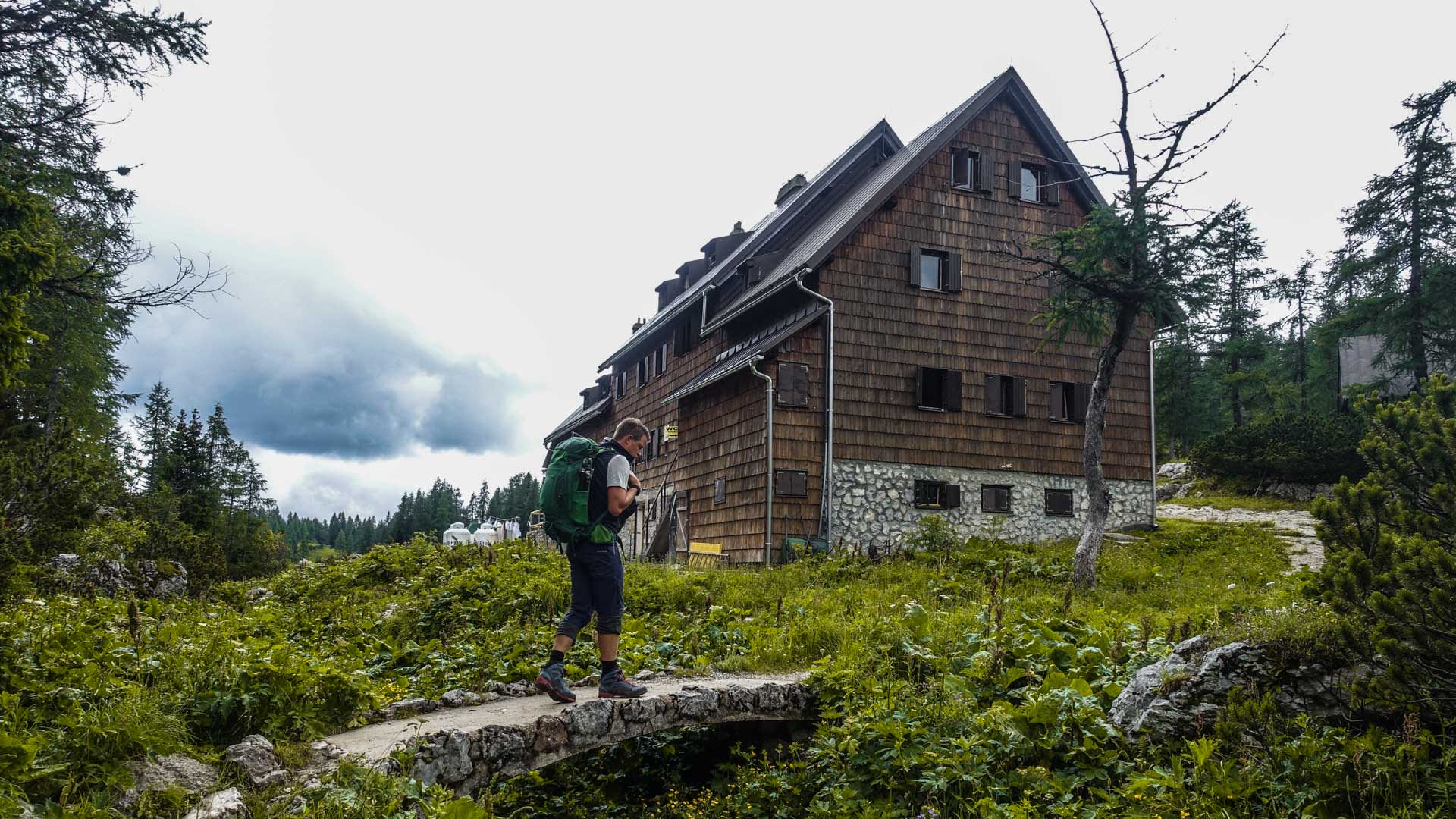 Bergwanderung zur Sieben-Seen-Hütte: Bergwanderer mit Rucksack überquert Steinbrücke vor der Holzschindel-Berghütte in Slowenien.
