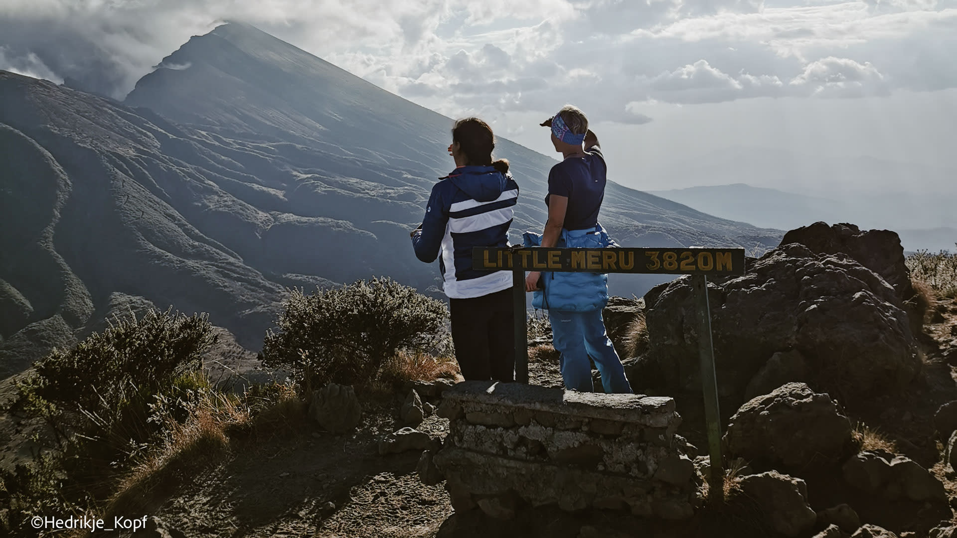 Zwei Höhenbergsteiger stehen am Schild des Little Meru 3820 m mit Blick auf den Vulkankegel des Mount Meru.
