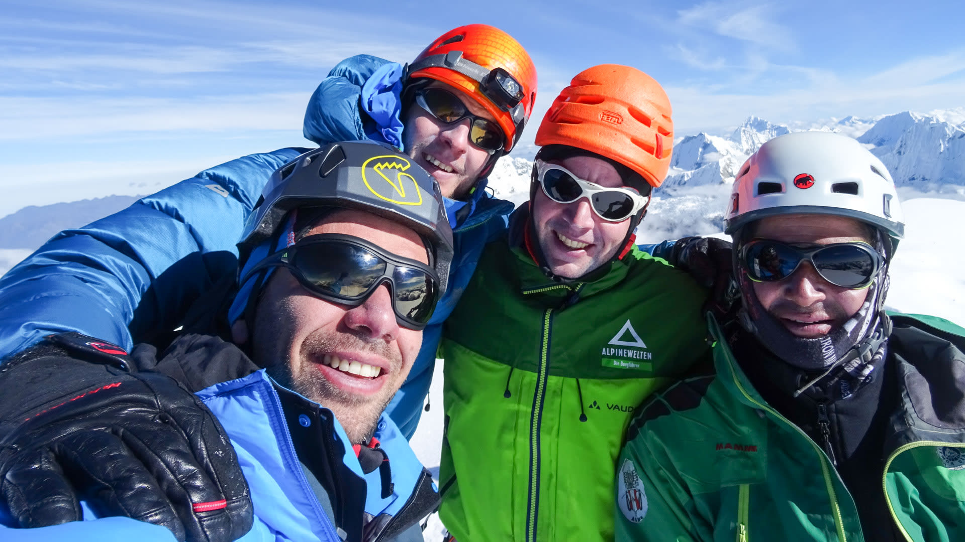 Expedition am Chopicalqui: vier Höhenbergsteiger mit Helm und Skibrille beim Gipfelfoto vor verschneiten Hochgebirgsgipfeln.