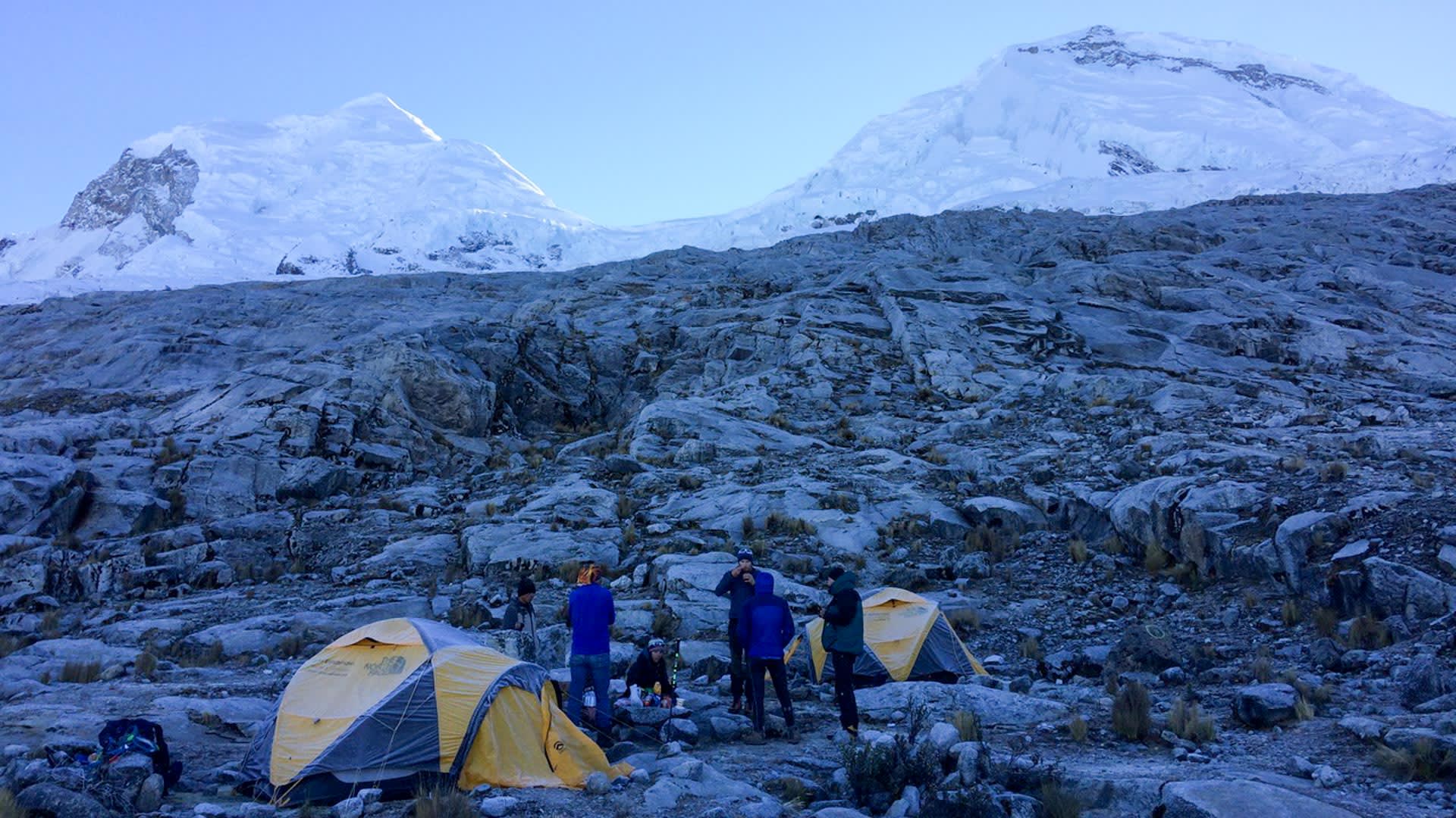 Expedition Huascarán: Höhenbergsteiger im Moränenlager bei Morgendämmerung, vergletscherte Gipfel im Hintergrund, Peru.