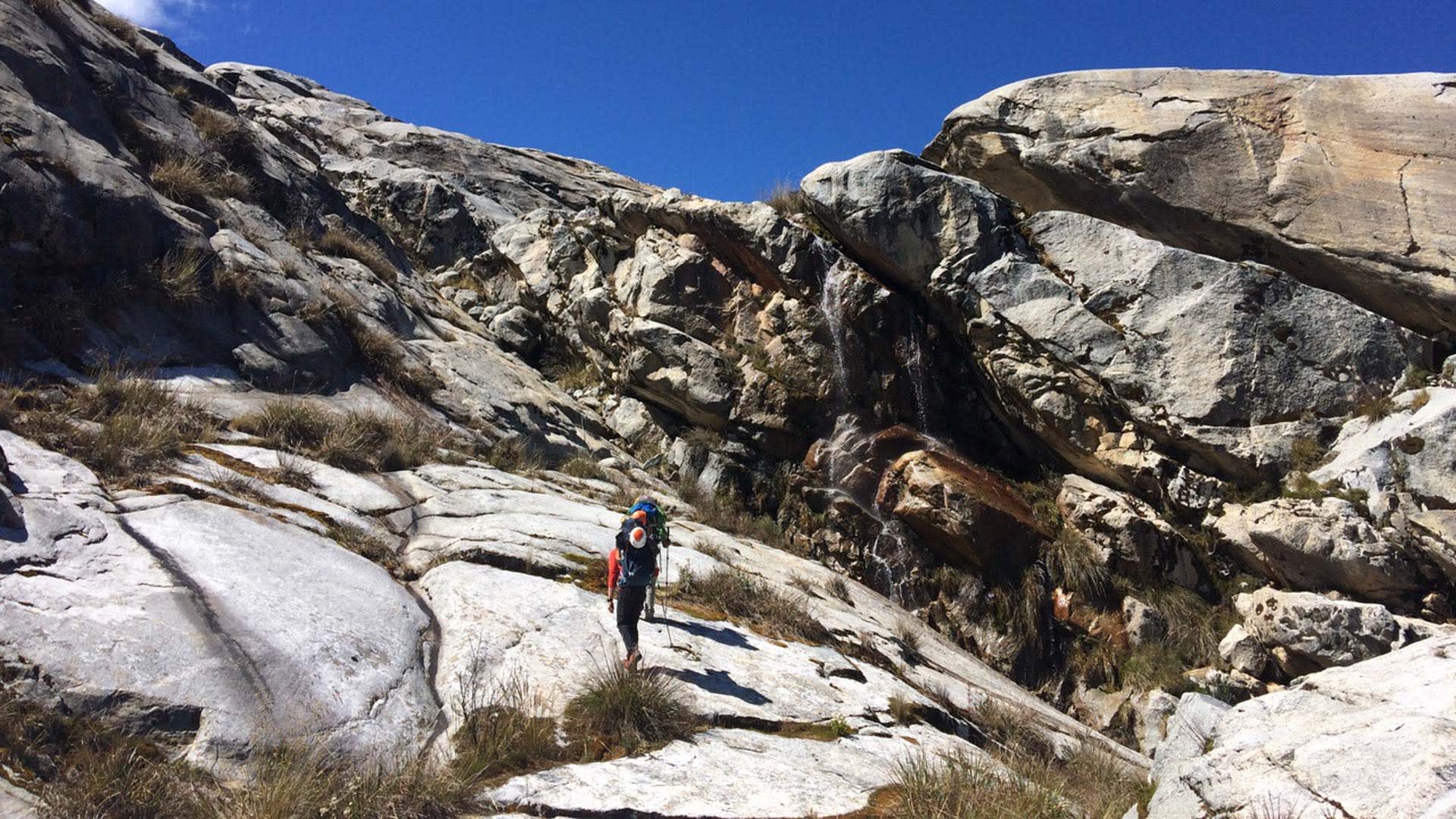Expedition Huascarán: Höhenbergsteiger mit blauem Rucksack und Wanderstock im Zustieg über Granitfelsen in Peru.