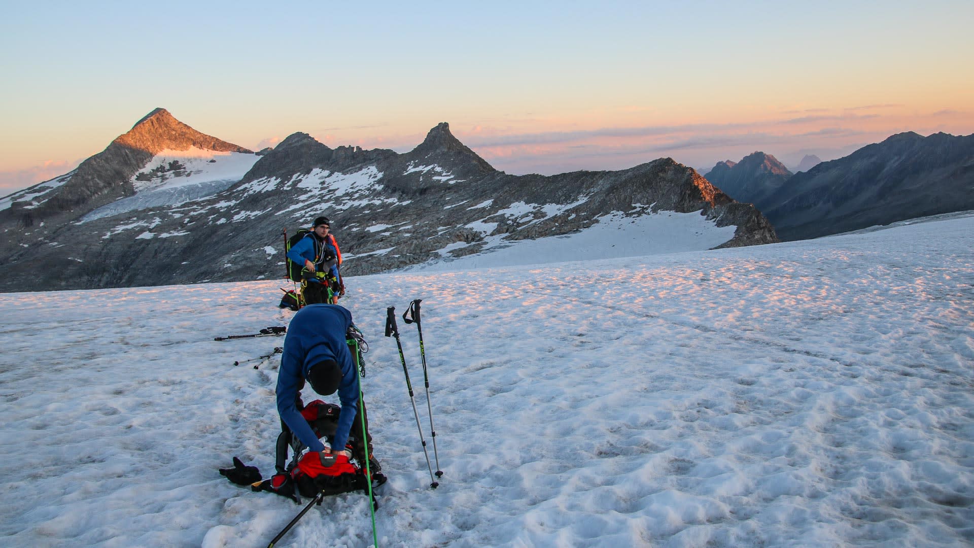 Zwei Bergsteiger auf einem Firnfeld am Großvenediger packen Ausrüstung bei Morgenlicht, Bergpanorama im Hintergrund.