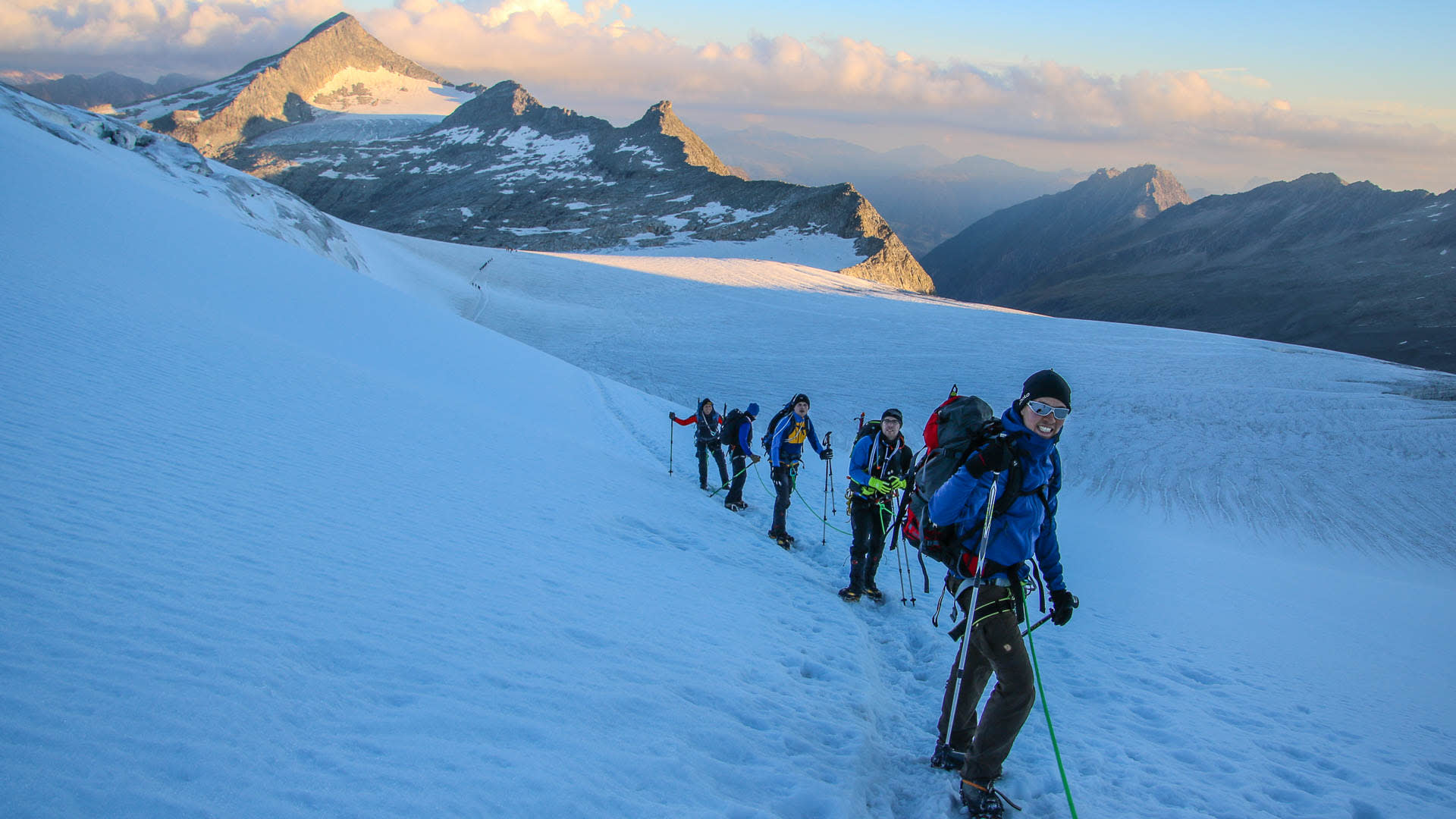 Fünf Bergsteiger mit Steigeisen und Rucksäcken steigen im Morgen- oder Abendlicht einen Schneehang zum Großvenediger auf.