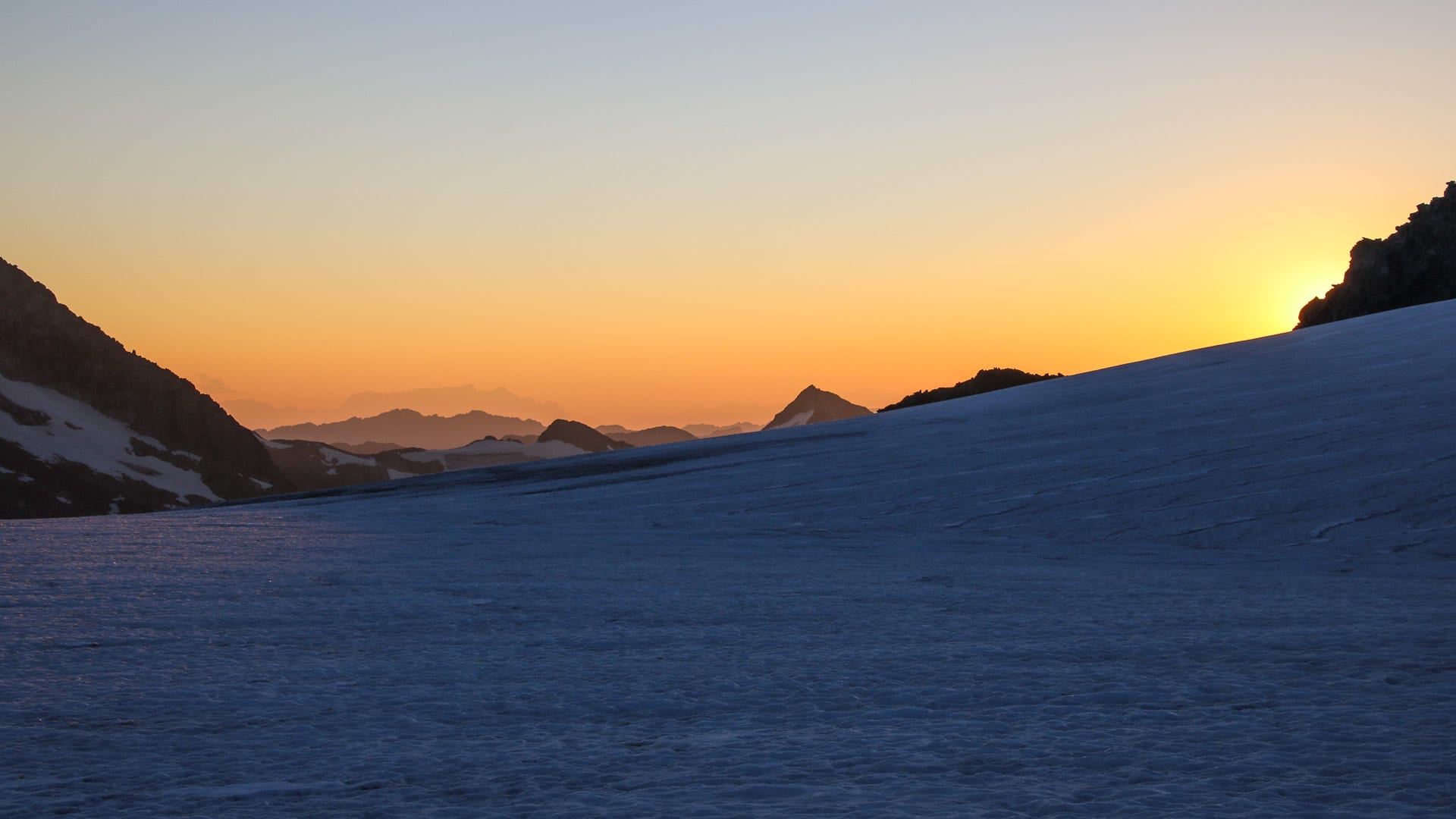 Schneebedeckter Hang am Großvenediger bei Sonnenaufgang mit orangegelbem Lichtschein und Bergsilhouetten im Hintergrund.