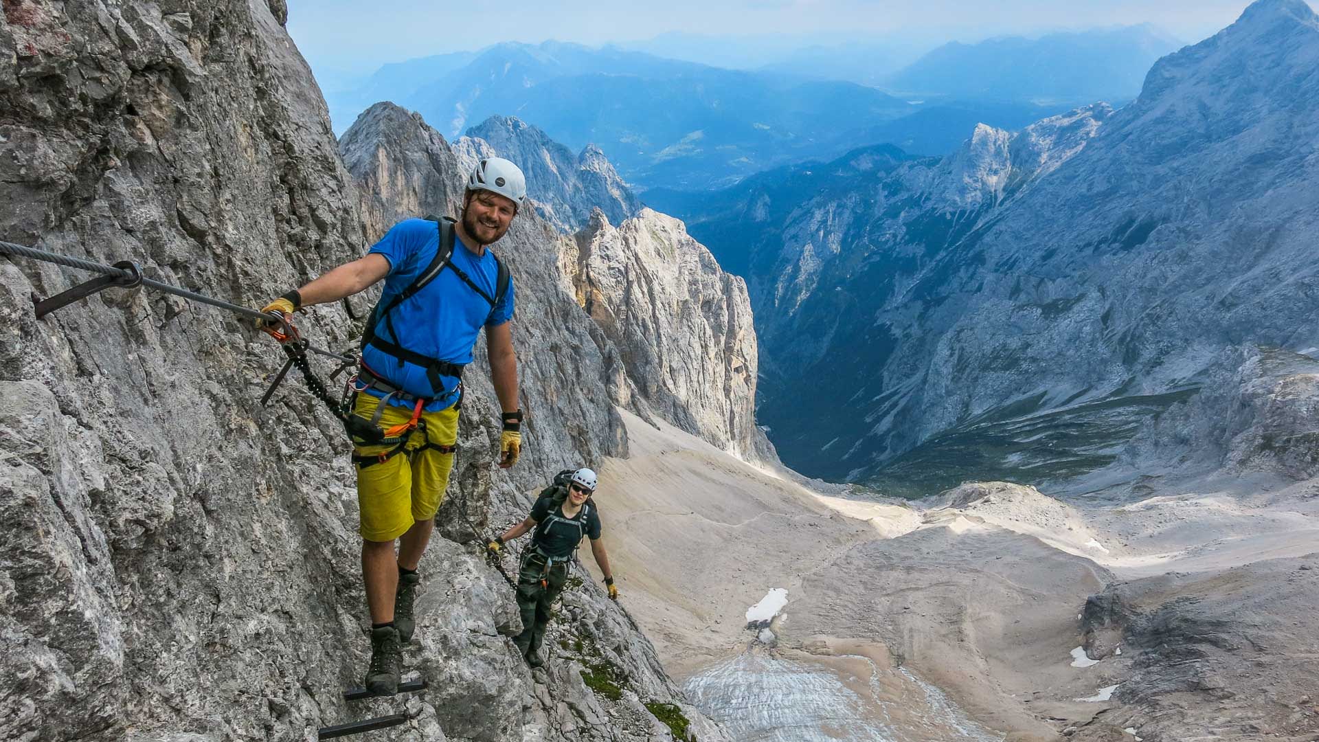 Zwei Klettersteiggeher an Eisenbügeln im Höllental, im Hintergrund der Gletscherrest und das Zugspitzmassiv.