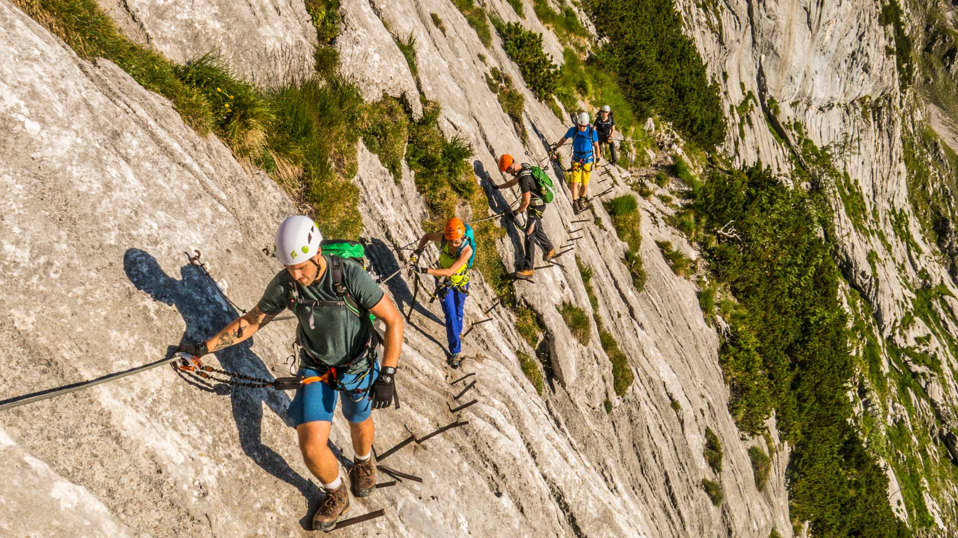 Fünf Klettersteiggeher traversieren an Eisenstiften und Fixseil eine senkrechte Kalkfelswand am Klettersteig zur Zugspitze.