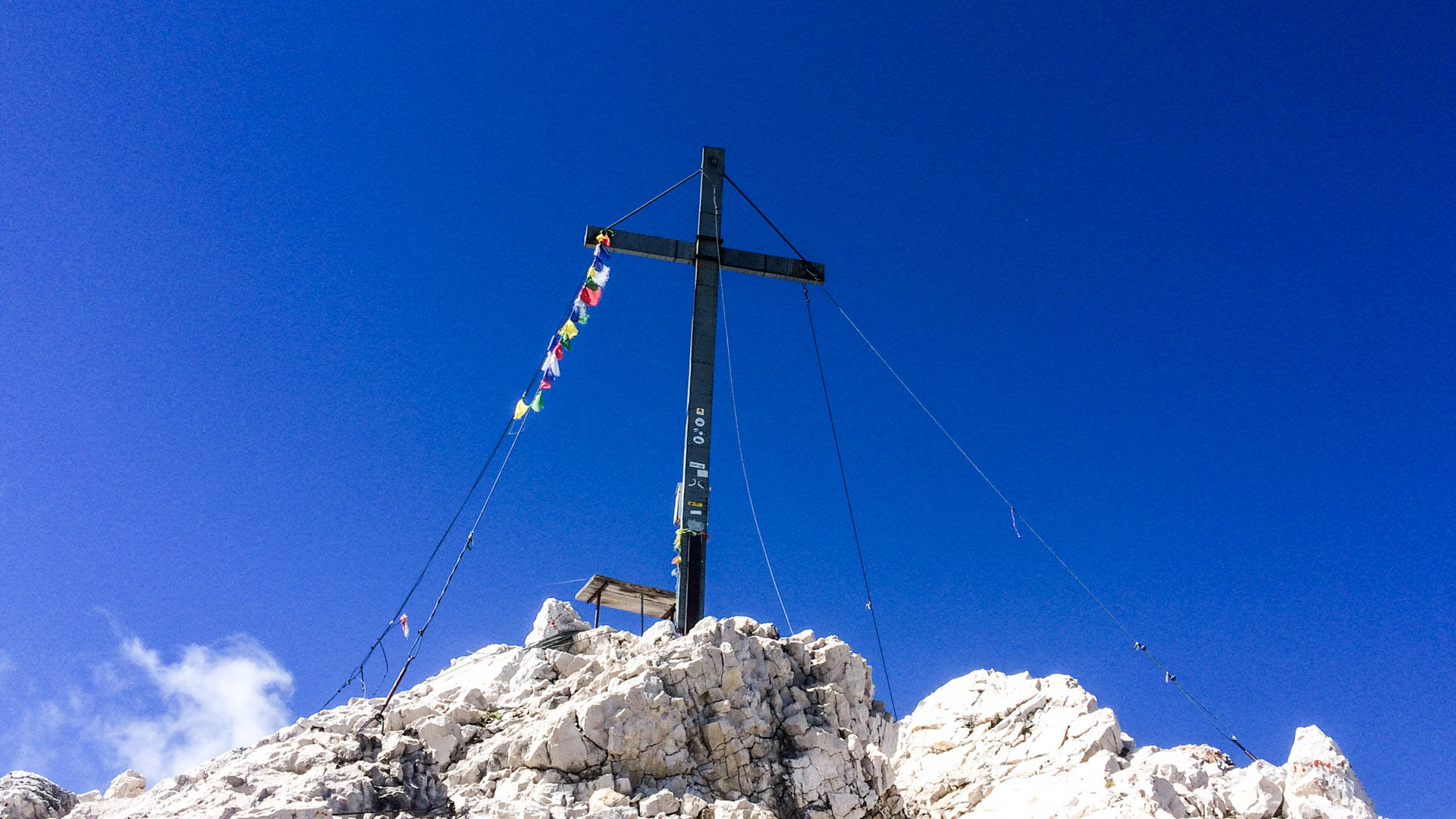 Klettersteig Alpspitze: Metallgipfelkreuz mit Gebetsfahnen auf hellem Kalksteingipfel vor wolkenlosem Himmel.