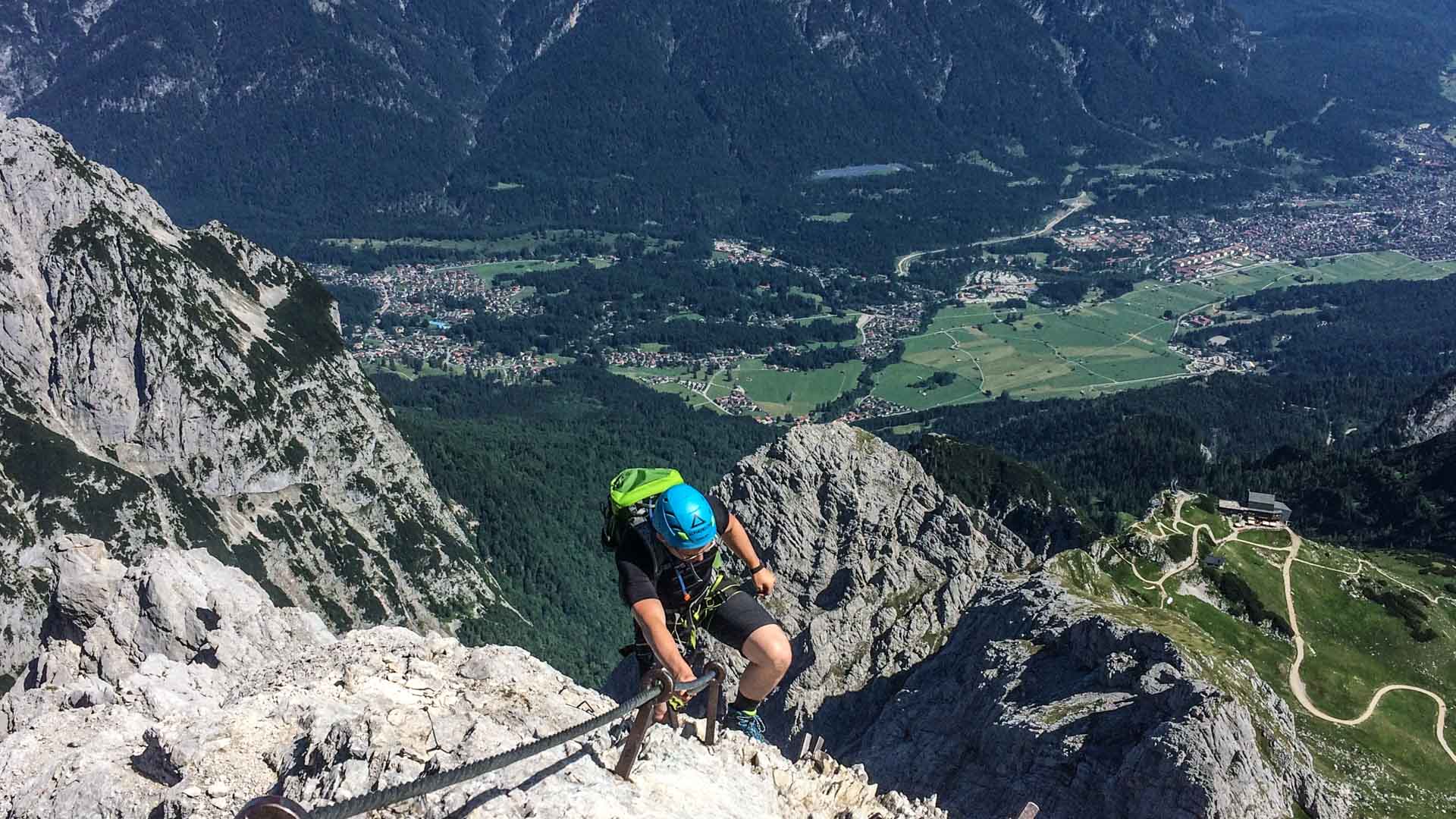 Klettersteig an der Alpspitze: Klettersteiggeher greift an Eisenklammern einer Kalkfelswand, Tal im Hintergrund.