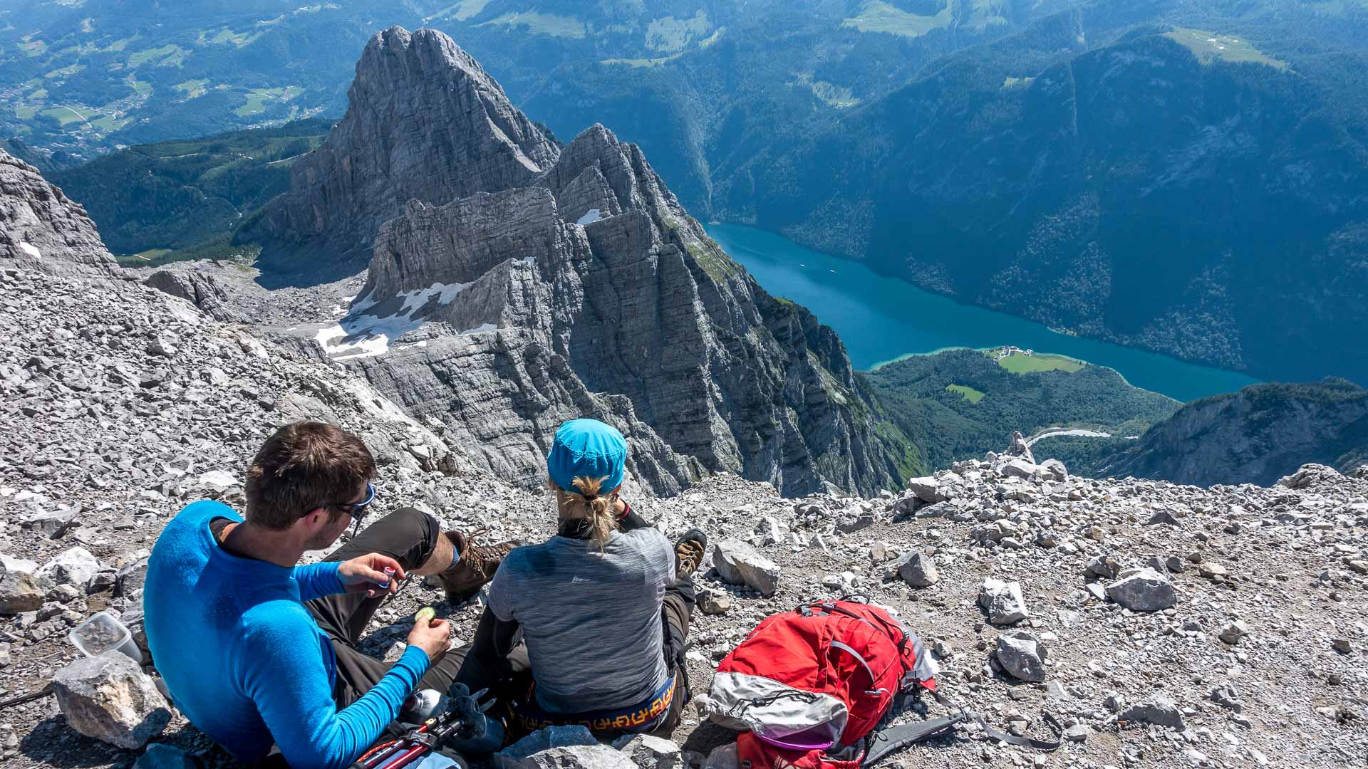 Klettersteig-Pause auf dem Watzmanngrat: zwei Personen rasten auf Geröll mit grandiosem Ausblick auf St. Bartholomäe im Tal.