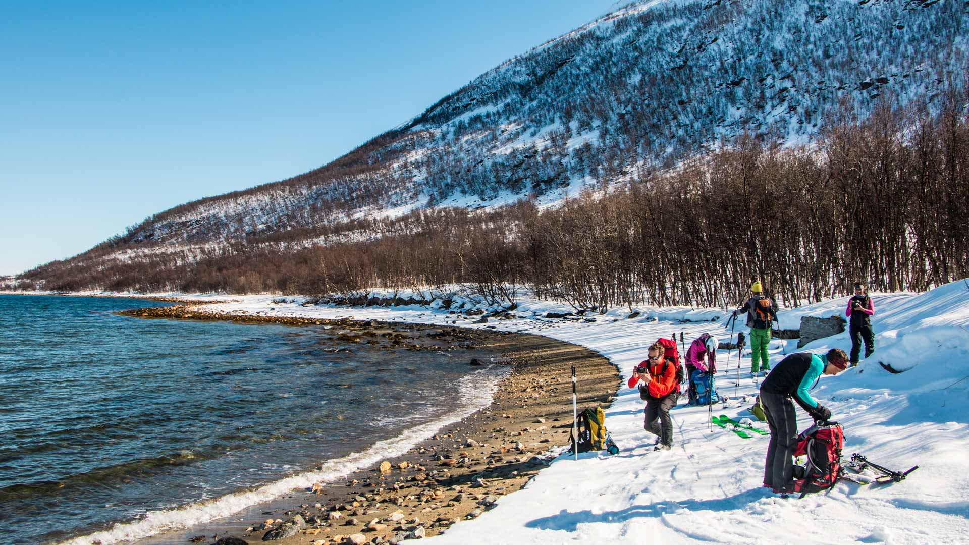 Fünf Skitourengeher rasten mit Tourenskiern und Rucksäcken am Ufer des Lyngenfjords in Norwegen.