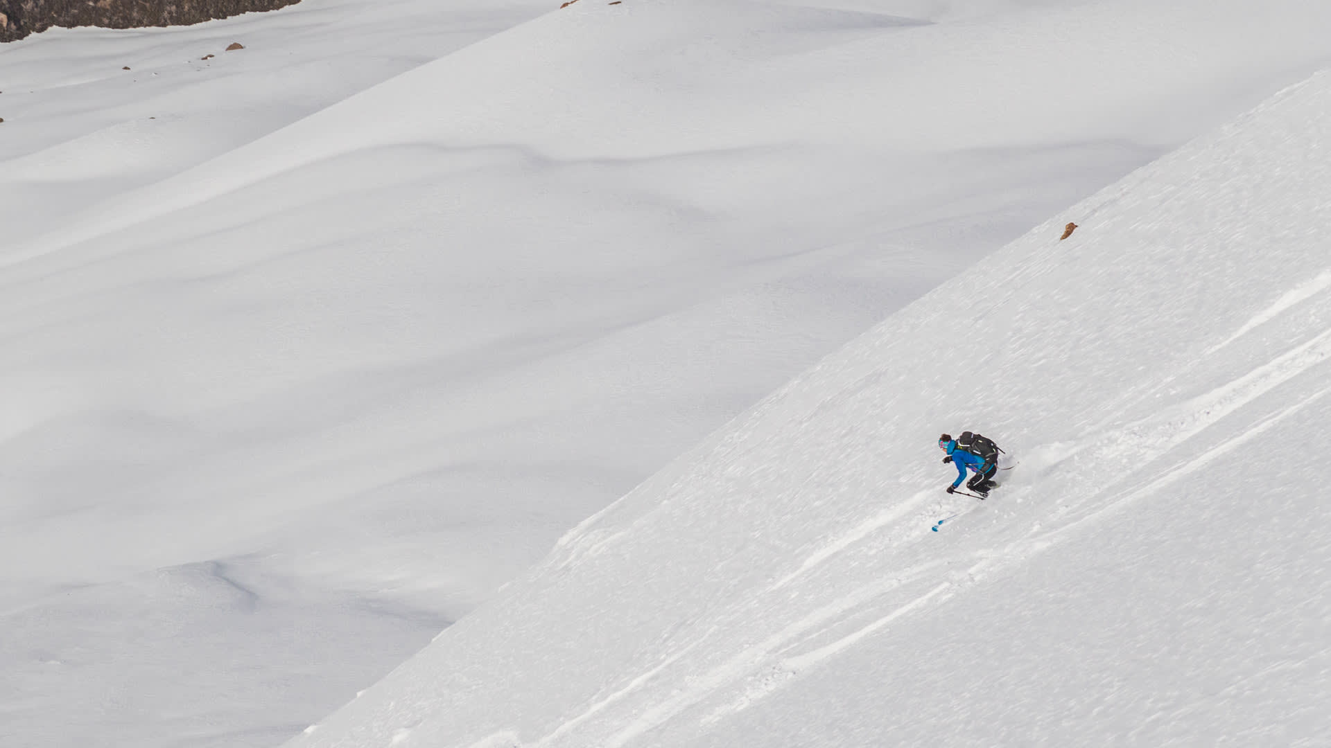 Skitour-Abfahrt in Kirgistan: Skitourengeher mit blauer Jacke und Helm fährt einen steilen schneebedeckten Hang hinunter.