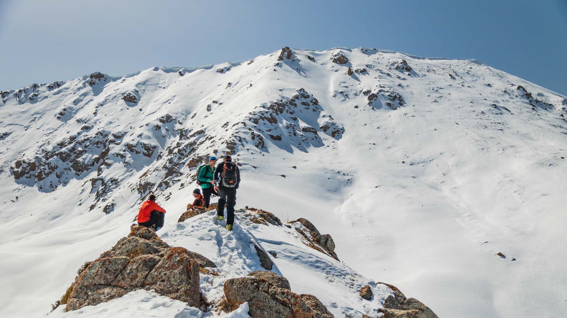Vier Skitourengeher pausieren auf felsigem Bergkamm in Kirgisistan, schneebedeckter Gipfel und blauer Himmel im Hintergrund.