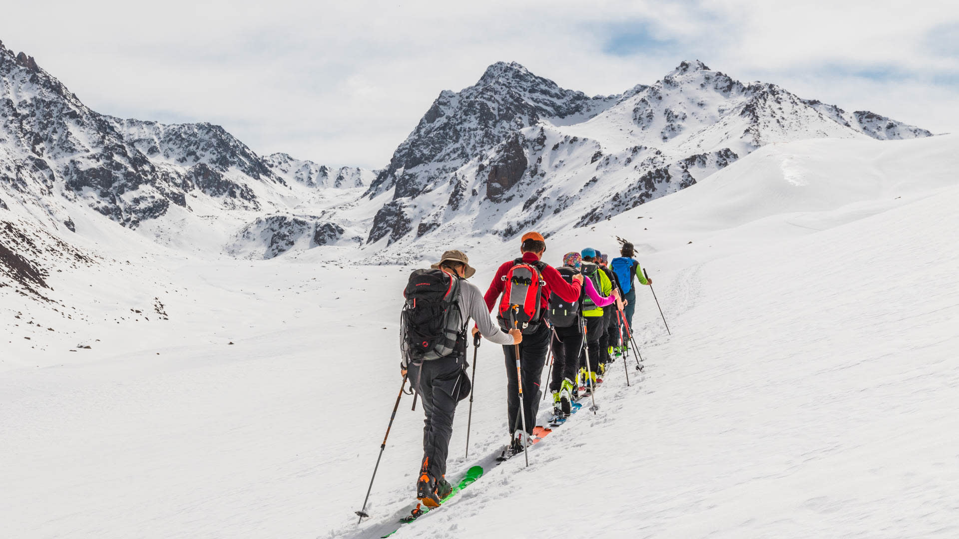 Sieben Skitourengeher steigen in einer Reihe mit Fellen am Ski durch ein offenes Schneebecken in Kirgisien auf.