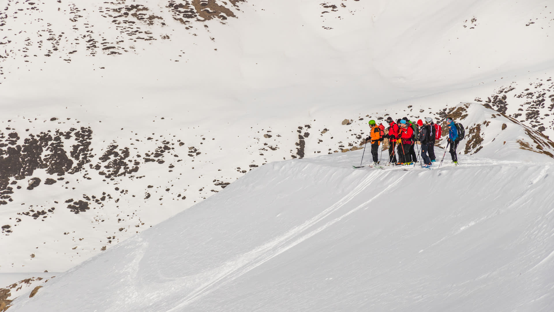 Skitour in Kirgistan: Gruppe von sieben Skitourengehern mit Fellen pausiert auf einem Schneehang vor felsigen Berggipfeln.