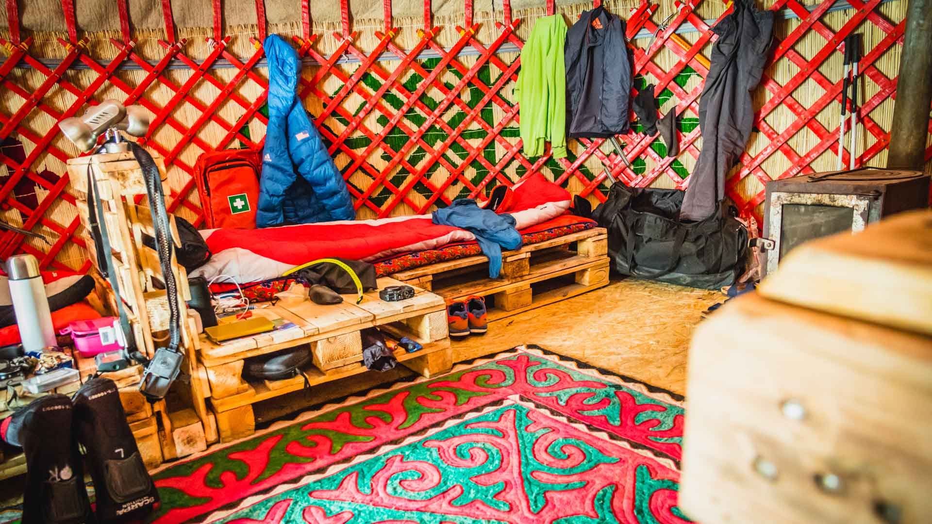 Skitour in Kirgistan: Innenansicht einer Jurte mit Schlafpaletten, Ausrüstung und traditionellem Holzofen als Unterkunft.