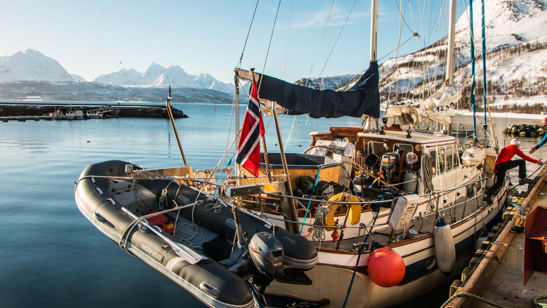 Skitour Lyngenalpen: Segelboot mit norwegischer Flagge und Schlauchboot am Kai, im Hintergrund schneebedeckte Gipfel.