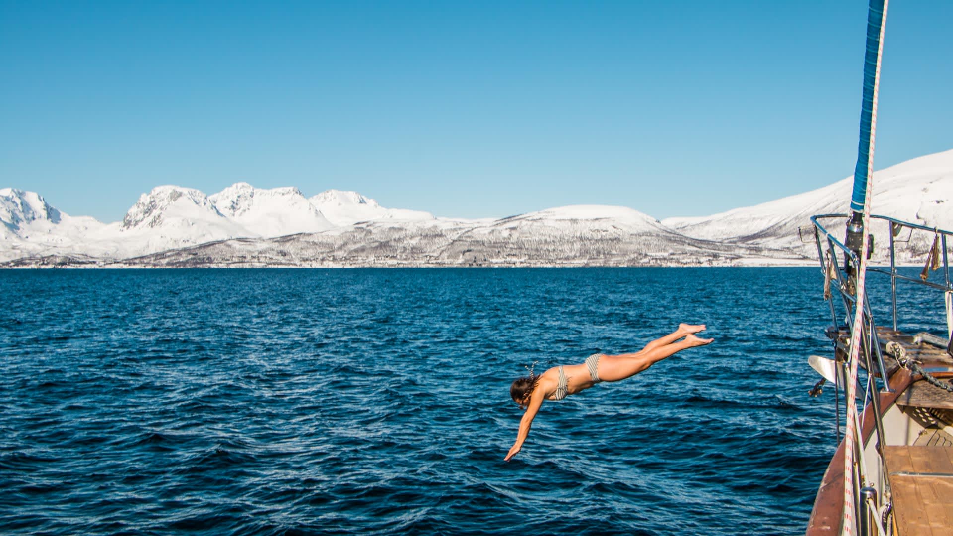 Nach der Skitour in Lyngen springt eine Person im Bikini vom Deck des Segelboots in den blauen Fjord.