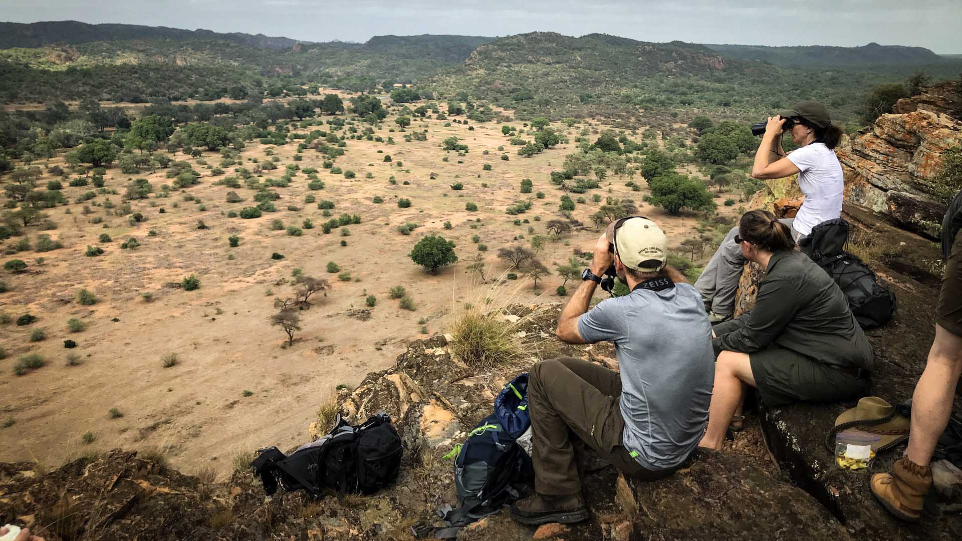 Trekking in Südafrika: vier Trekker rasten auf Felsvorsprung mit Fernglas über weiter Savannenlandschaft.