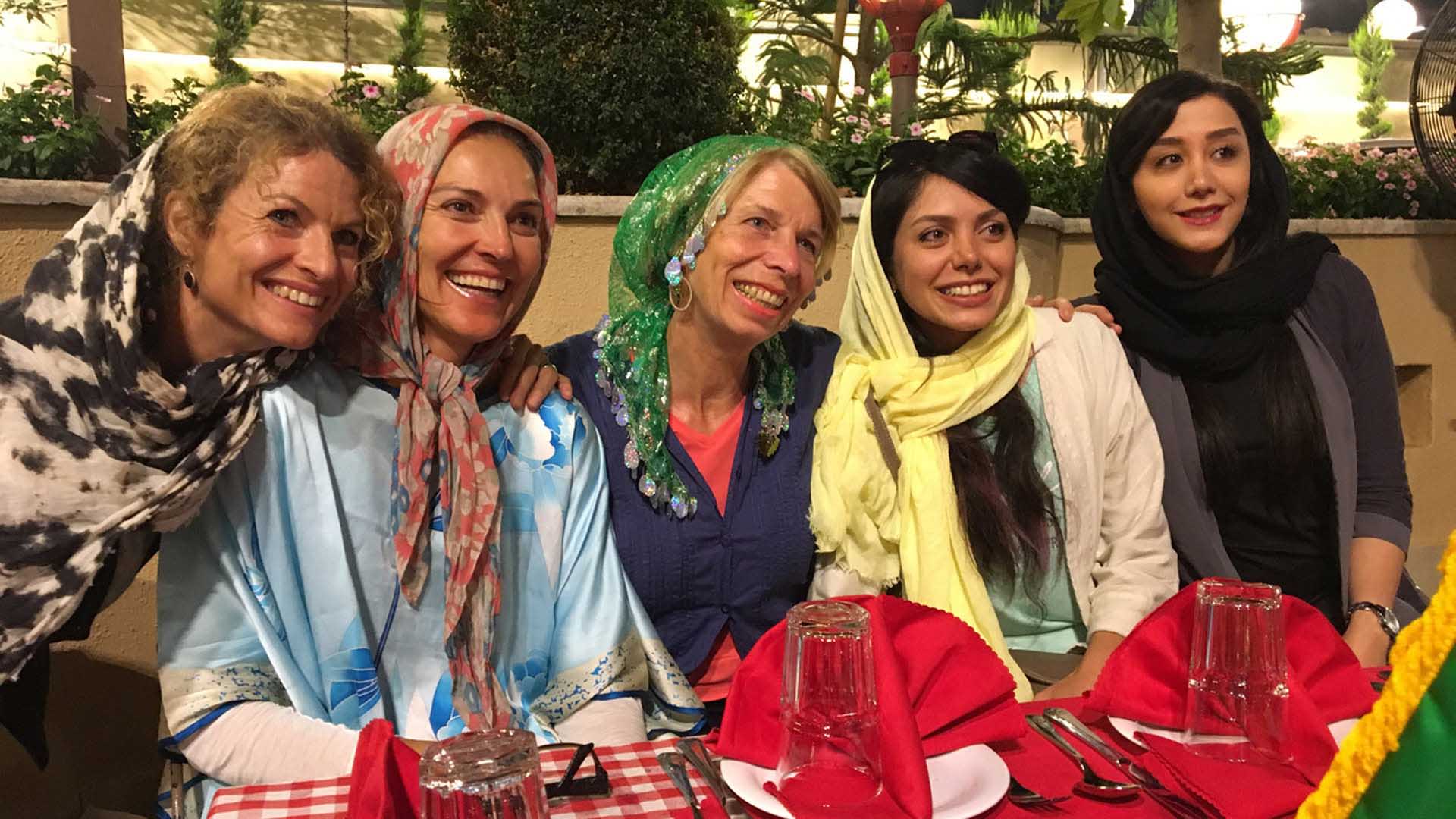 Trekking im Iran: Fünf Frauen mit Kopftüchern sitzen lächelnd an gedecktem Tisch auf einer begrünten Restaurantterrasse im Iran.