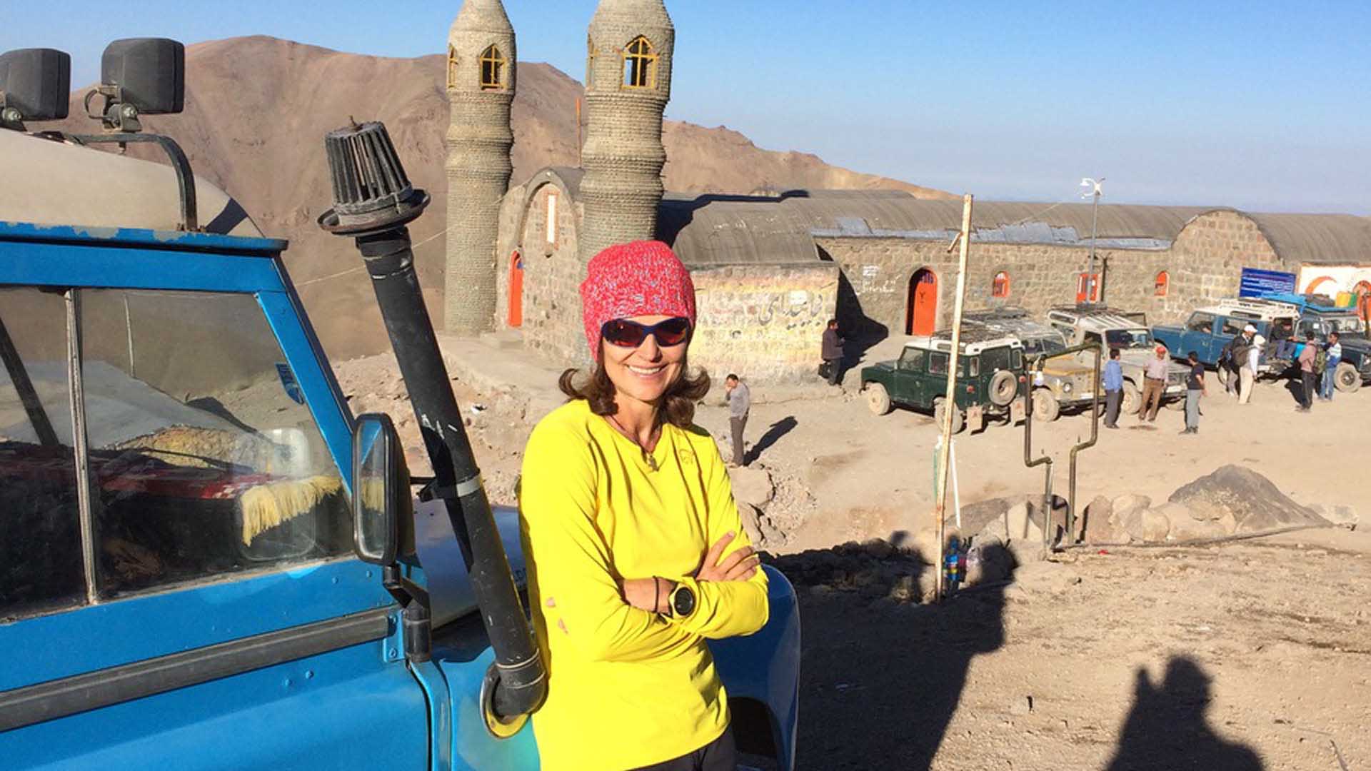 Trekking im Iran: Frau in gelber Jacke lehnt an blauem Geländefahrzeug vor einem Steingebäude mit geflochtenen Minarett-Türmen