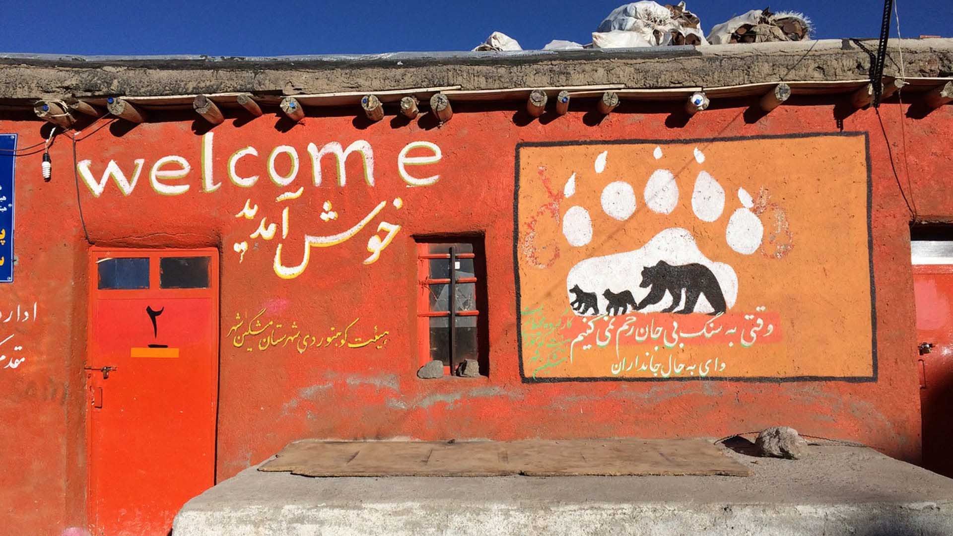 Trekking im Iran: Orangerote Lehmfassade mit weißem Welcome-Schriftzug, persischer Inschrift und Bären-Wandbild unter blauem Himmel.