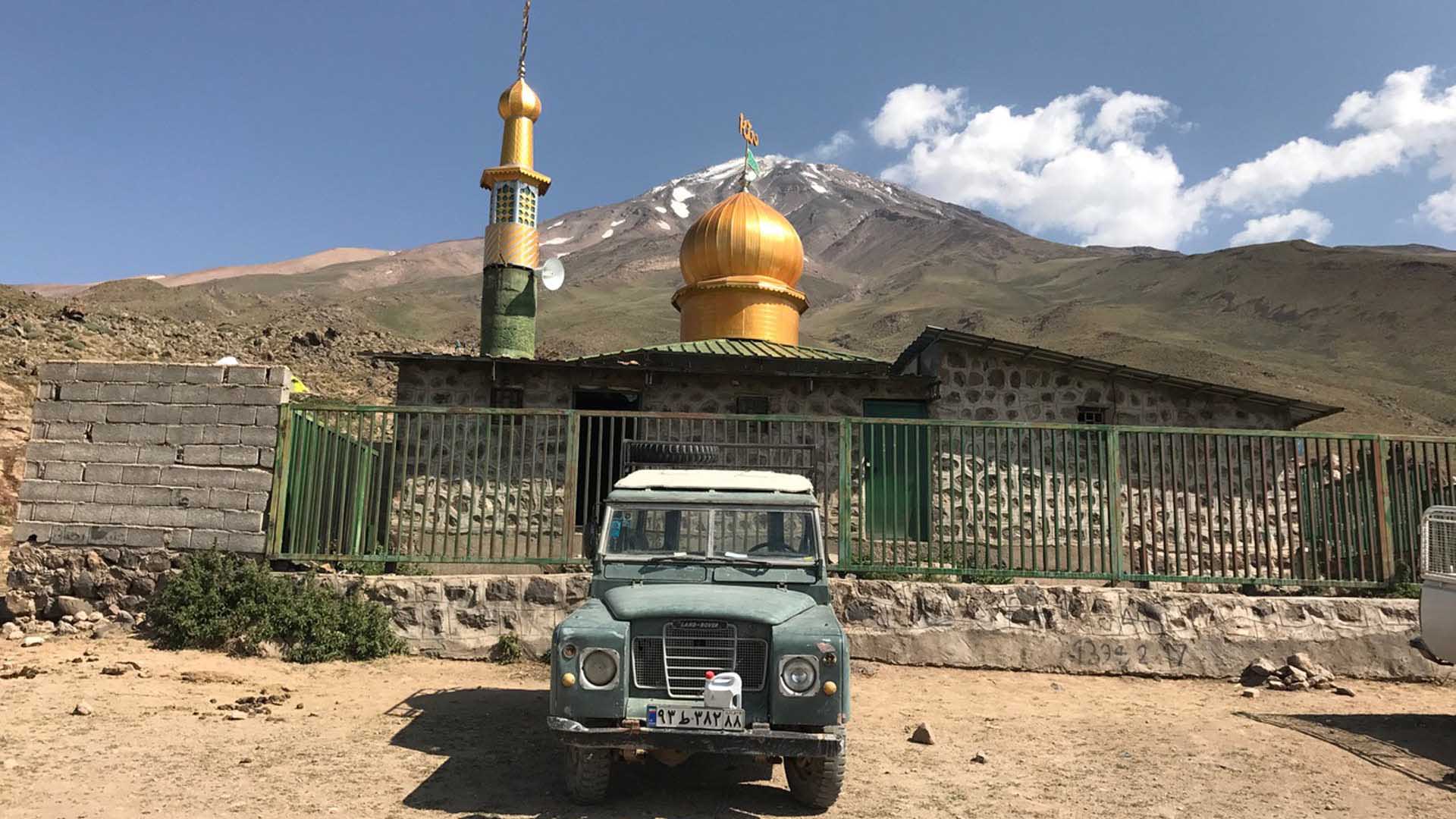 Trekking im Iran: Land Rover vor kleiner Moschee mit goldenen Kuppeln und schneebedecktem Vulkankegel im Hintergrund.