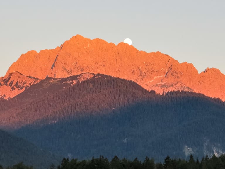 Bergwanderung im Karwendel: Felsiger Gipfelgrat im Alpenglühen mit Mondscheibe knapp über dem Kamm.