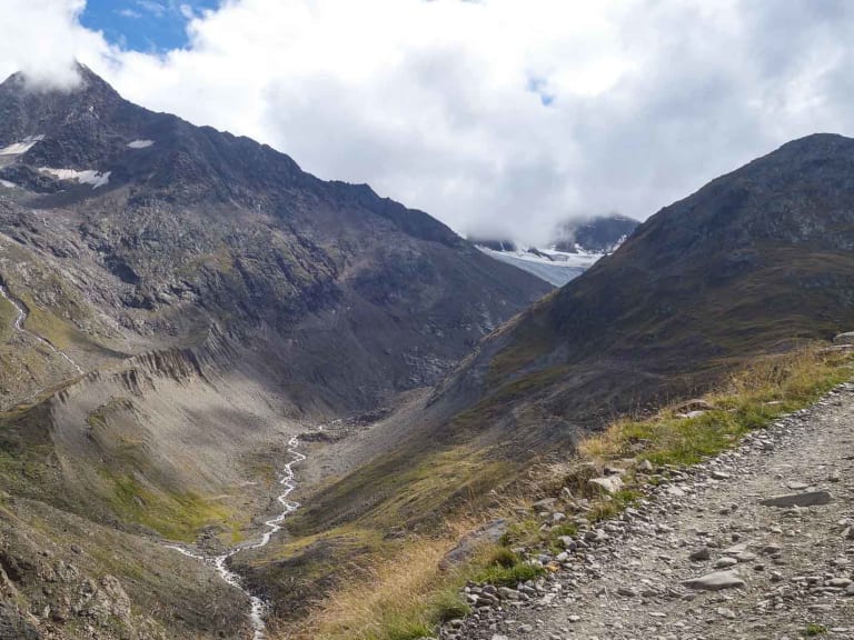 Bergwanderung in der Bernina: Schotterweg durch karges Hochtal mit mäanderndem Gletscherbach und Gletscherzungen im Hintergrund.