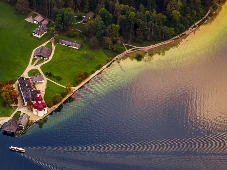Bergwanderung am Königssee: Luftaufnahme der Halbinsel St. Bartholomä mit rotem Zwiebelturm, Holzsteg und Ausflugboot auf dem See.