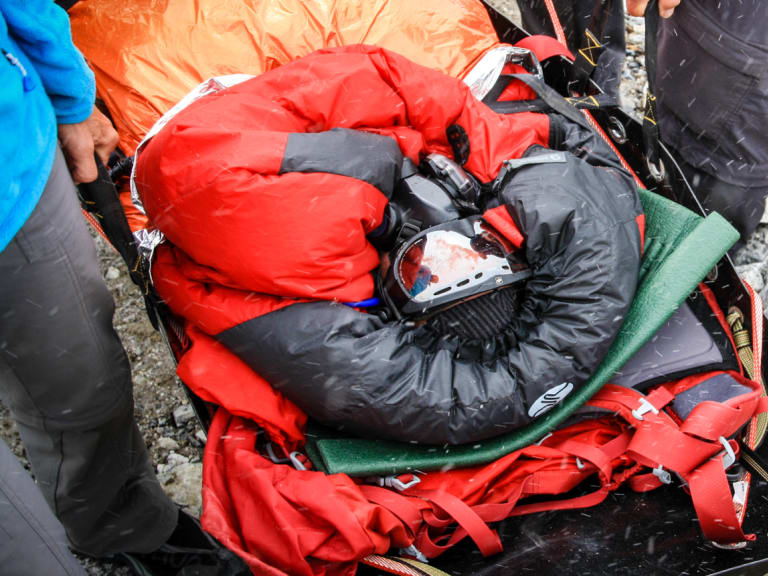 Expedition-Rettung am 8000er: Höhenbergsteiger auf roter Akia-Trage fixiert, zwei Helfer auf Felsblöcken bei Schneefall.