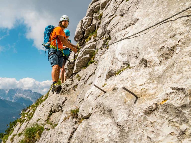 Zwei Kletterer traversieren ein Klettersteig-Drahtseil an einer steilen Kalkfelswand in den Berchtesgadener Alpen mit Bergpanorama