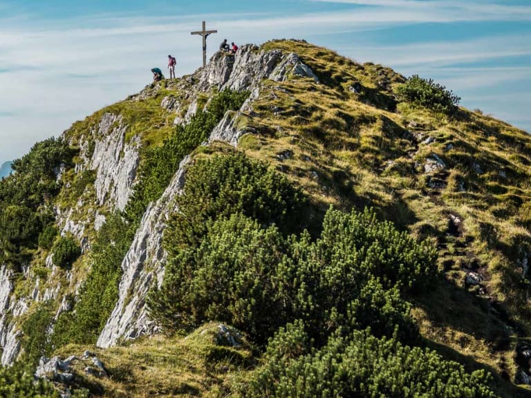 Drei Klettersteiggeher am Gipfelkreuz des Untersberg mit dem Watzmann-Massiv im dunstigen Hintergrund.