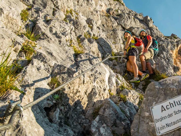 Zwei Klettersteiggeher mit Gurten stehen vor dem Warnschild Beginn der Schwierigkeiten am Untersberg-Klettersteig.