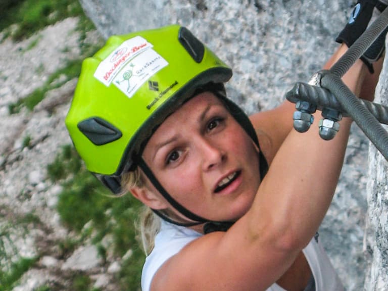 Klettersteiggeherin greift an Stahlbügel einer senkrechten Kalkfelsenwand am Staufen, gelber Helm.