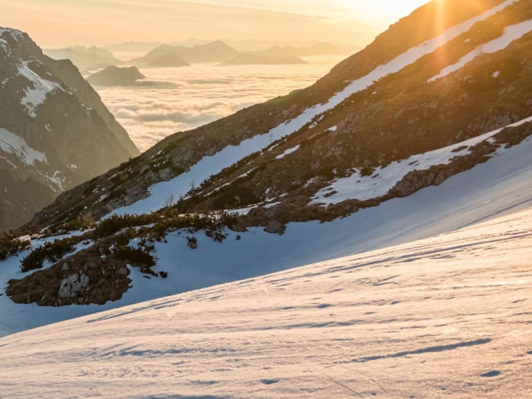 Drei Skitourengeher steigen diagonal einen Schneehang in den Berchtesgadener Alpen auf, im Hintergrund steile Felsgipfel