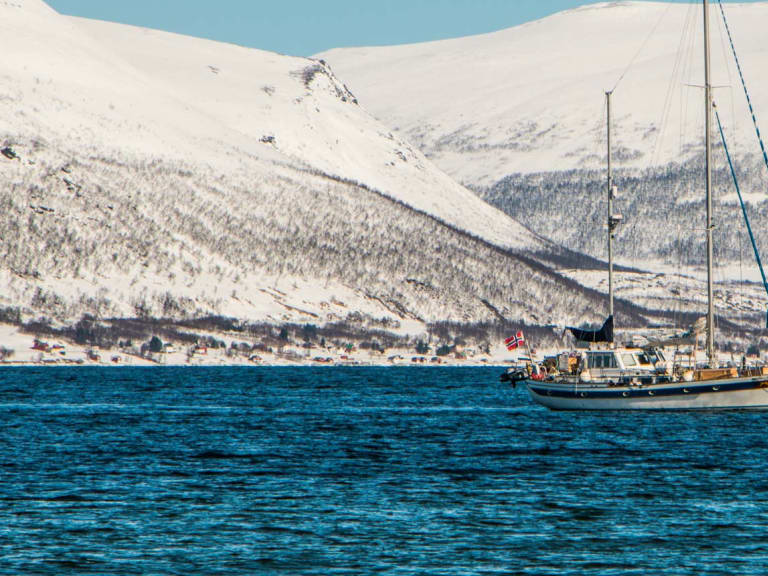 Skitour in Norwegen: Segelboot mit norwegischer Flagge auf dem Fjord bei Tromsø, dahinter schneebedeckte Berghänge